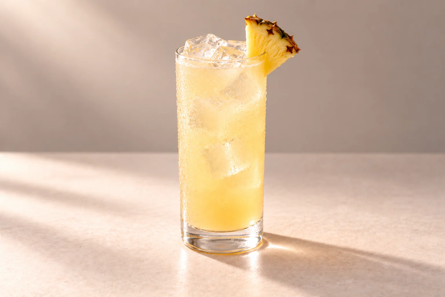 Jasmine Tropical Collins made with Lychee, Coconut & Jasmine Chakra Vodka, served over ice in a Collins glass with a pineapple wedge on an ivory surface.