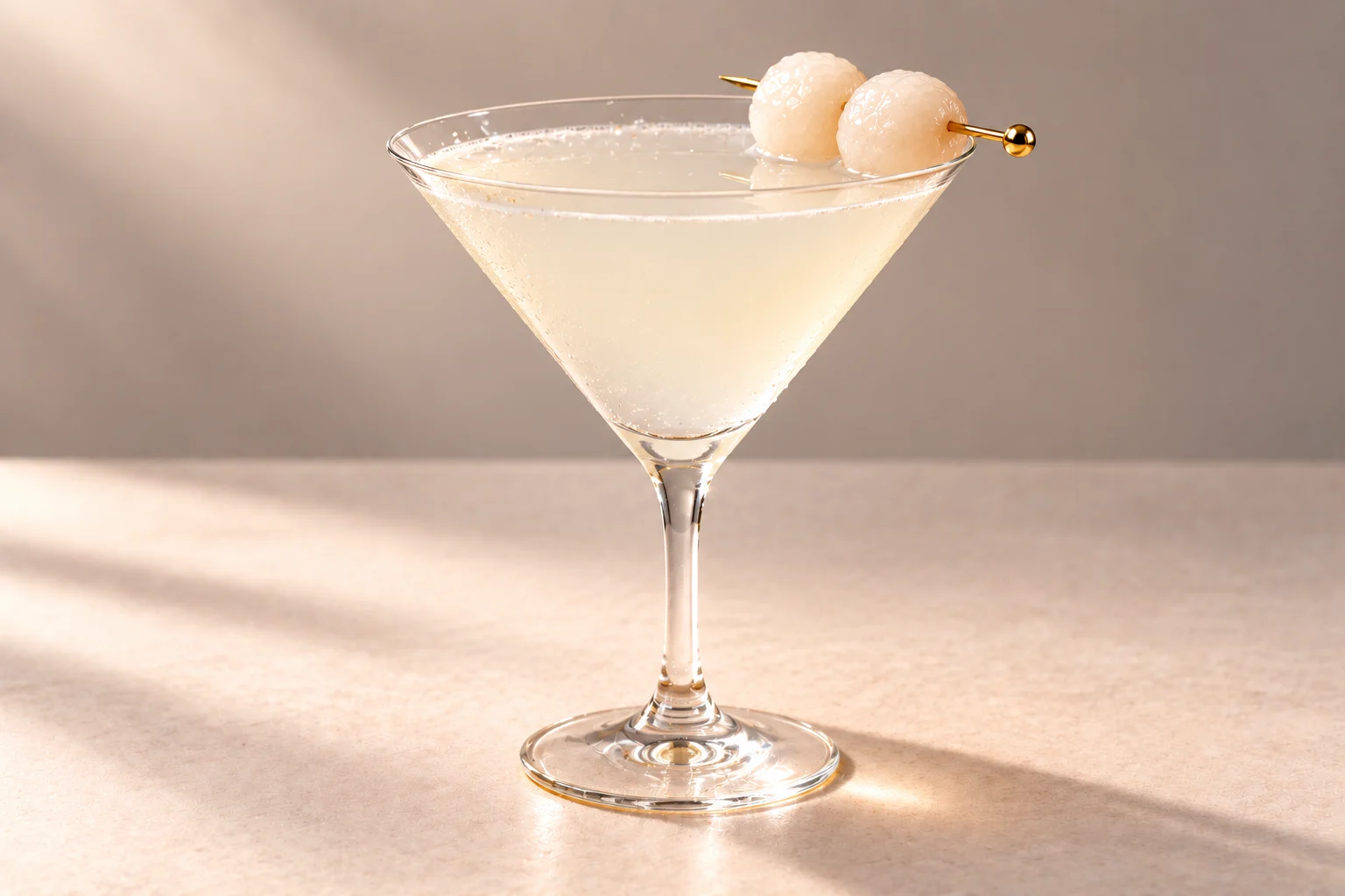 Coconut Lychee Martini made with Lychee, Coconut & Jasmine Chakra Vodka, served in a martini glass with two lychees on a gold cocktail pick on an ivory surface.
