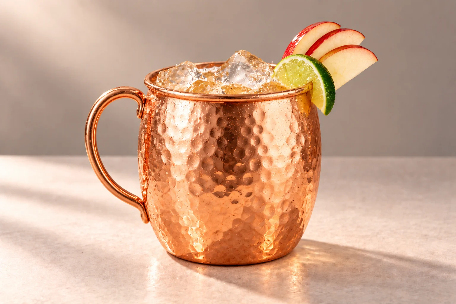 Red Apple & Ginger Mule made with Red Apple & Ginger Root Chakra Vodka, served over ice in a hammered copper mug with apple slices and a lime wedge on an ivory surface.