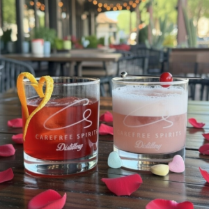 Valentine’s Day special cocktail at Carefree Spirit Distillery: vibrant red-pink drink with heart-shaped orange twist garnish, served in branded rocks glass over ice with scattered red rose petals