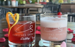 Valentine’s Day special cocktail at Carefree Spirit Distillery: vibrant red-pink drink with heart-shaped orange twist garnish, served in branded rocks glass over ice with scattered red rose petals